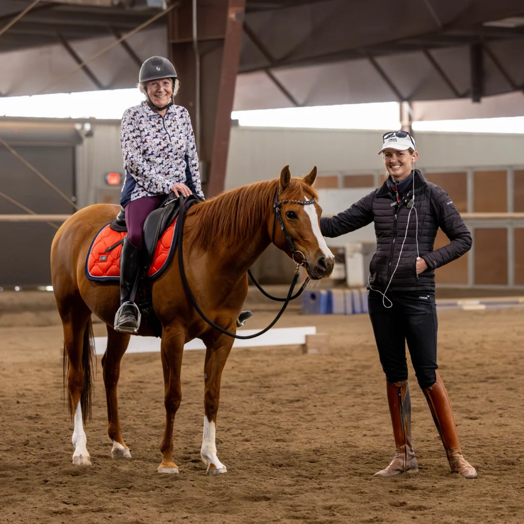 A smiling person wearing a helmet sits on a saddled chestnut horse inside an indoor arena, while another smiling person stands beside them, resting a hand on the horse. Both are dressed in riding attire.