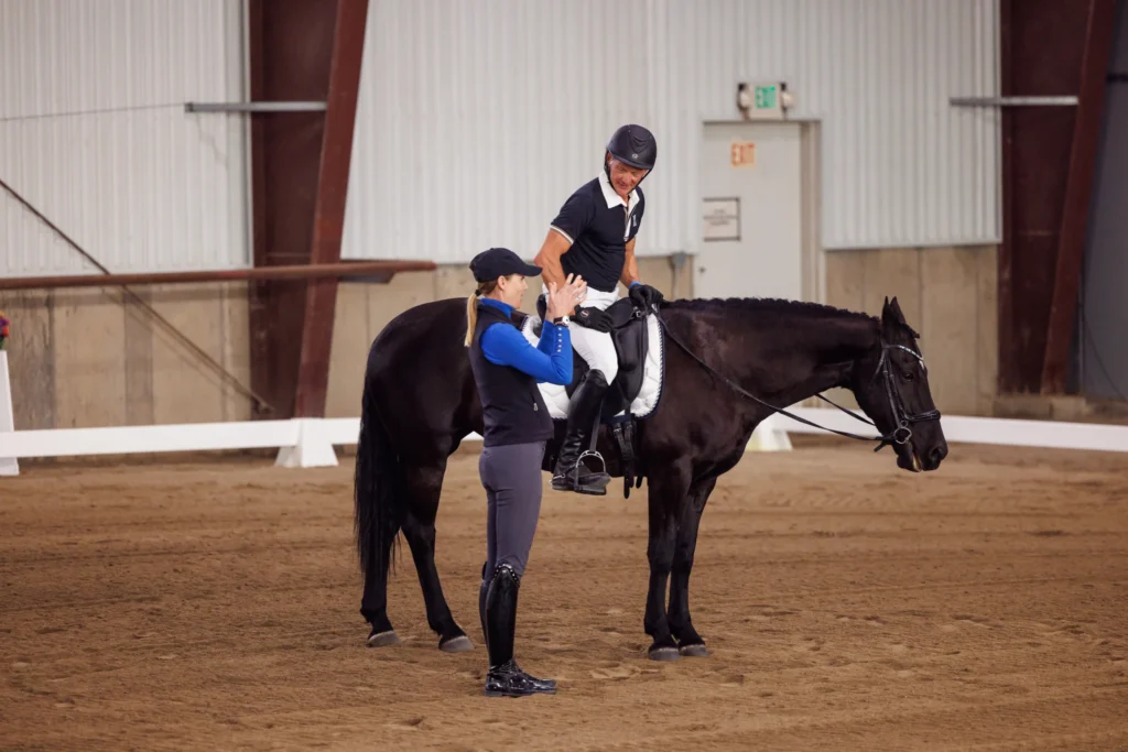 A rider on a bay horse performs a collected halt and extends their hand for a high five with another individual standing at their side in an indoor arena. Both individuals are outfitted in casual riding gear rather than formal dressage attire, echoing the encouraging and collaborative spirit characteristic of Amelia Newcomb’s training environment.