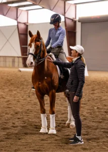 A person on horseback listens to trainer Amelia Newcomb indoors. The horse has white markings and leg wraps. The rider wears a helmet; the trainer gestures while watching closely. Both are dressed in equestrian attire.