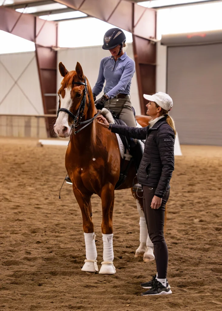 A person on horseback listens to trainer Amelia Newcomb indoors. The horse has white markings and leg wraps. The rider wears a helmet; the trainer gestures while watching closely. Both are dressed in equestrian attire.