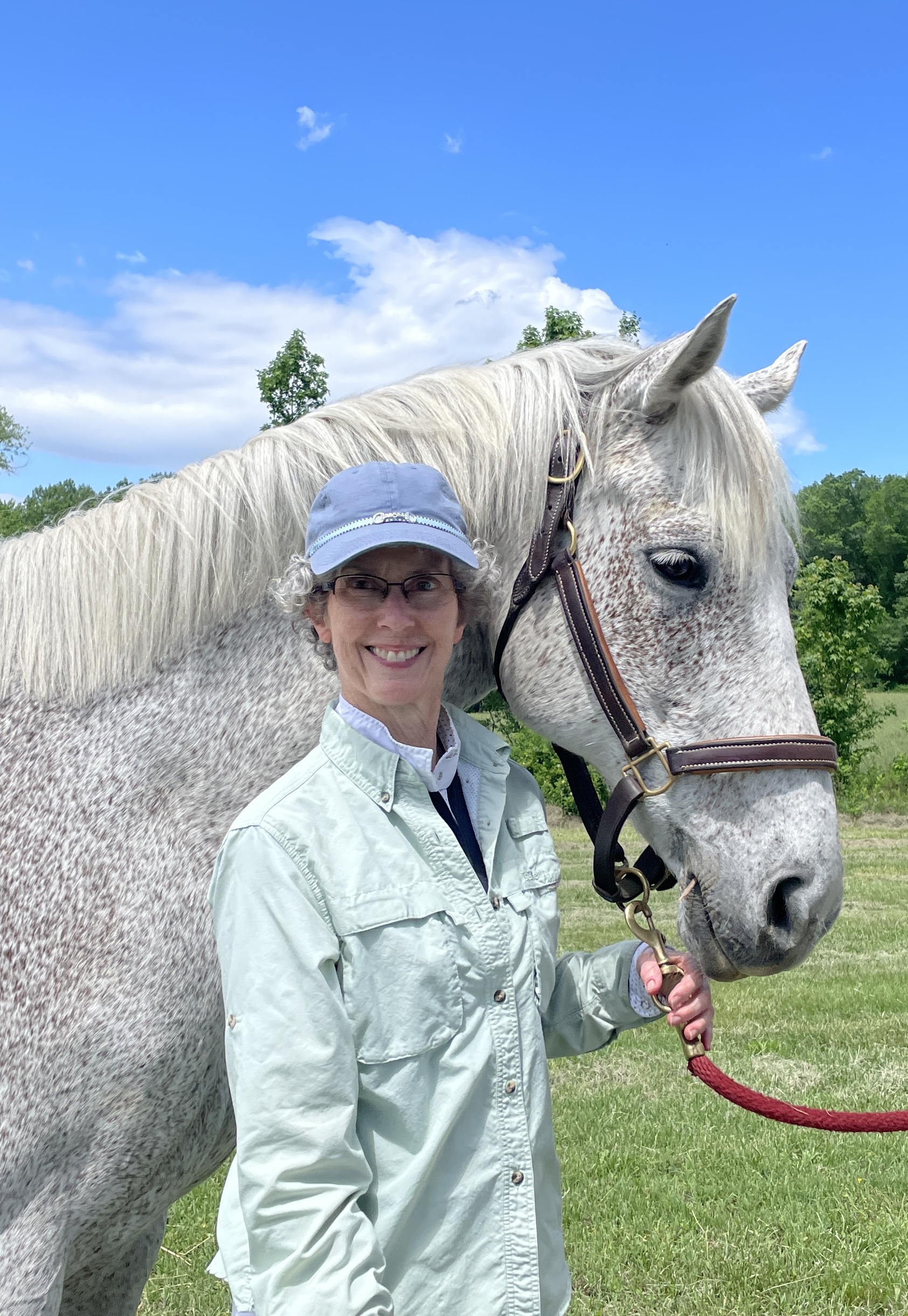 A smiling student in a light green shirt and blue cap stands outdoors, holding the lead rope of a grey horse on a sunny day; the setting features green trees and a clear blue sky in the background.