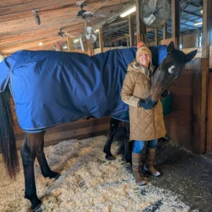 A woman in a brown winter coat and hat stands in a horse stable, smiling and holding the head of a large dark horse covered with a blue blanket. The stable has wooden walls and sawdust on the floor.