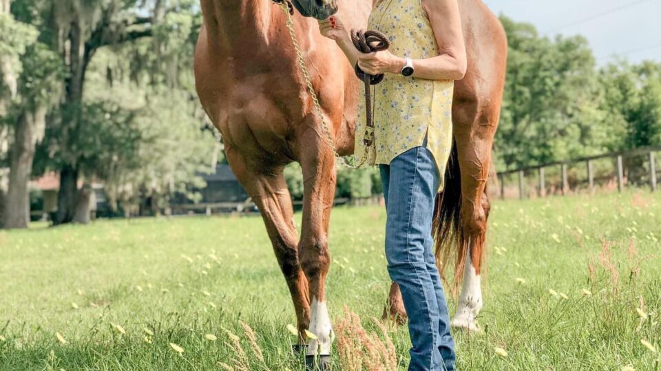 Strides Member Janet, wearing a straw hat, yellow sleeveless shirt, and blue jeans, stands outdoors in a grassy field, holding the reins of a brown horse. Trees and a clear sky form the background.