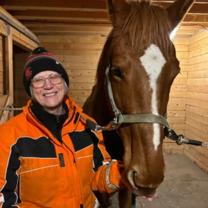 A smiling person in glasses and an orange jacket stands next to a brown horse with a white heart-shaped mark on its forehead inside a wooden stable.