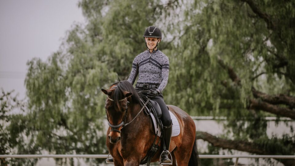 A person in equestrian gear, including a helmet and patterned long-sleeve shirt, rides a dark brown horse in an outdoor arena with trees in the background, as seen during a July 2023 event featured in the news.