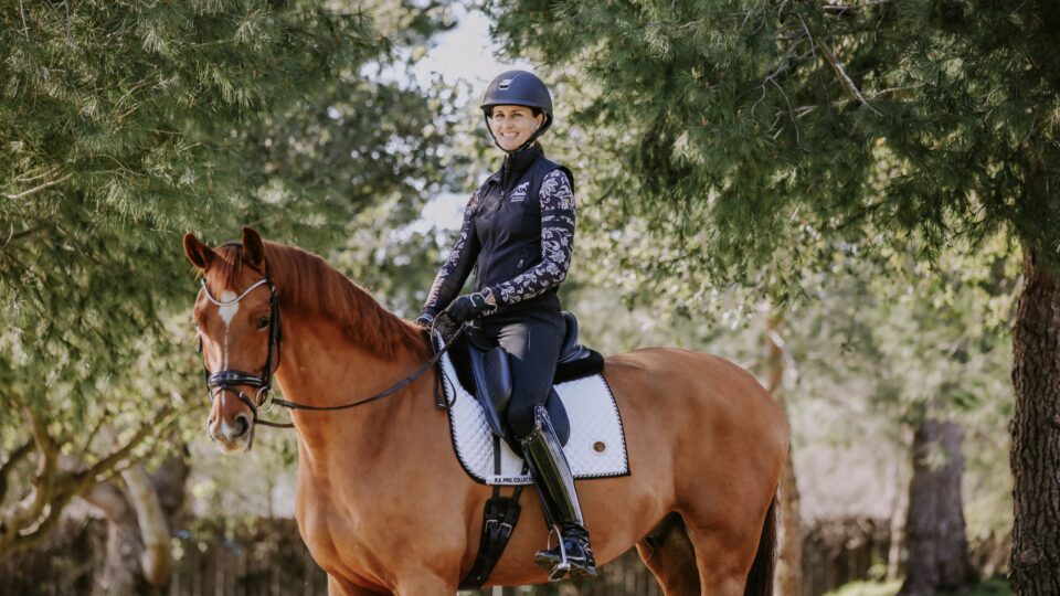 A person wearing a helmet and riding gear demonstrates excellent rider position while sitting on a brown horse in an outdoor Omaha setting surrounded by green trees and sunlight. Both appear calm and relaxed.