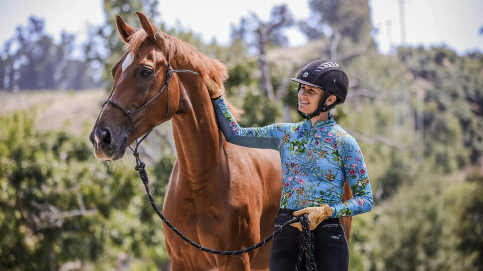 A person wearing a helmet and floral shirt smiles while standing beside a brown horse, holding its lead and gently touching its neck—a perfect moment showcasing horse training and the importance of good ground manners on a sunny day.