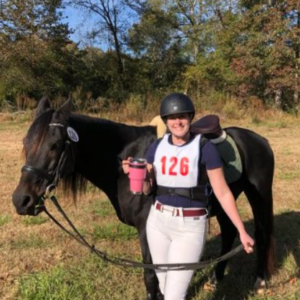A smiling person in riding gear and helmet, wearing a number 126 bib, stands outdoors holding a drink and leading a black horse by the reins, with autumn trees and grass in the background.