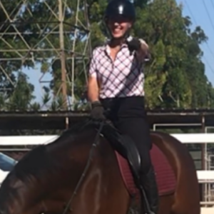 A person wearing a helmet and patterned shirt is smiling and pointing while sitting on a dark brown horse in an outdoor riding arena, perhaps inspired by tips from the Flying Changes Webinar. Trees, a fence, and a white car are visible in the background.