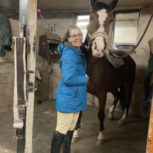A woman in a blue jacket and beige pants stands smiling next to a brown horse with a white stripe on its face inside a stable. The horse is wearing a halter and a blanket over its back.