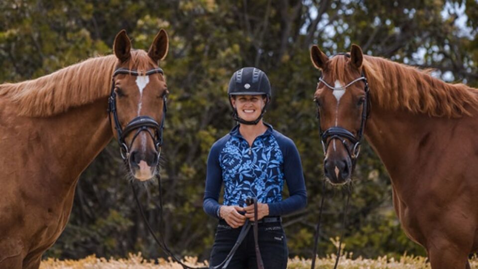 A person, featured in EuroDressage, wears a helmet and blue patterned shirt while smiling outdoors, holding the reins of two brown horses on either side. Trees and bushes fill the background.