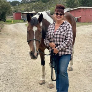 A woman wearing sunglasses and a plaid shirt stands smiling next to a brown and white horse on a gravel path, with stables and trees in the background.