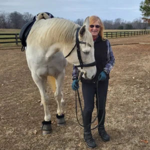 A woman wearing riding gear stands smiling beside a white horse, holding its reins in an outdoor paddock with a wooden fence and bare trees in the background.