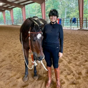 A person in riding gear and helmet stands next to a brown horse with a white blaze, inside an indoor riding arena with a dirt floor and metal beams. Trees are visible outside through the fencing.