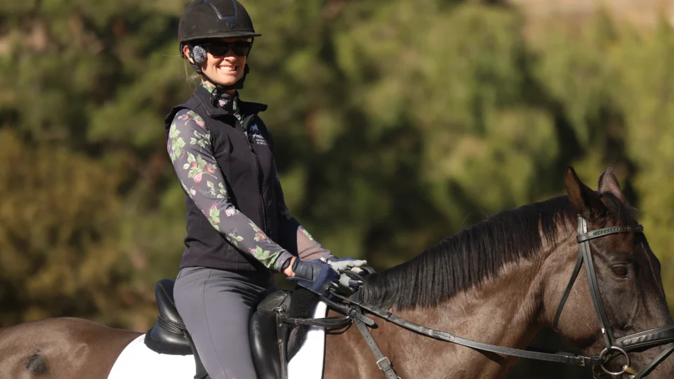 A person wearing a helmet, sunglasses, gloves, and a floral long-sleeve shirt rides a dark brown horse with a white saddle pad outdoors, surrounded by blurred green trees—perfect for listening to your favorite Confidence podcast.