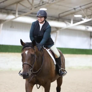 A person wearing a helmet, glasses, and riding gear smiles while sitting on a brown horse inside an indoor riding arena.