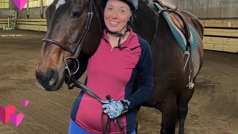 A smiling student in casual riding attire stands alongside Brandy, a dark bay horse, within an indoor arena. The image is adorned with pink and red heart graphics, lending a warm and supportive ambiance.