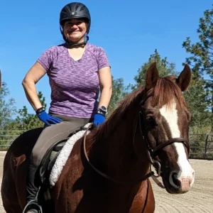 A woman wearing a helmet, purple shirt, gloves, and riding pants sits smiling on a brown horse with a white blaze, outdoors in a sandy arena with trees and a blue sky in the background.