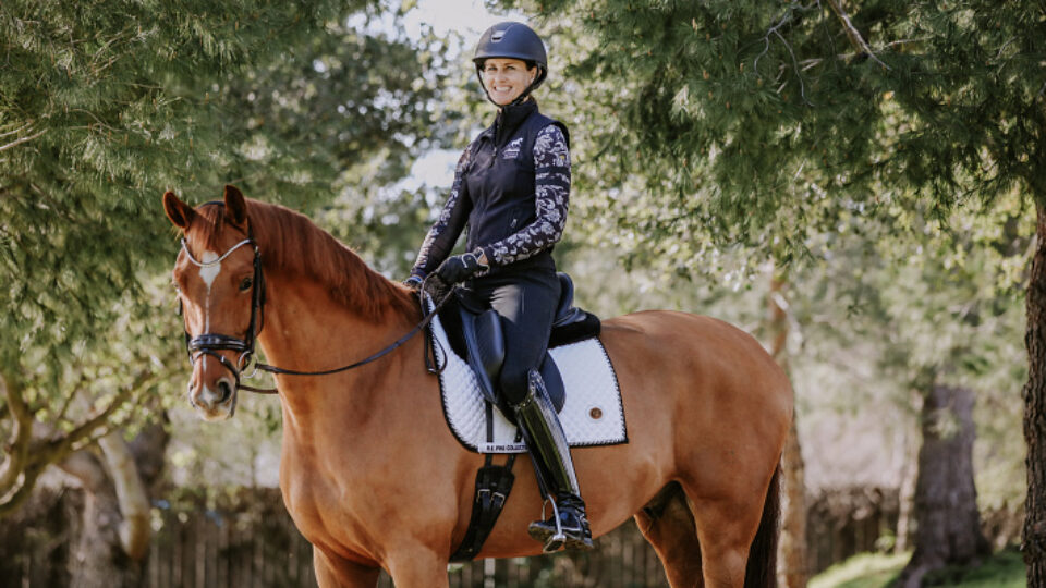 A person wearing riding gear and a helmet sits smiling on a brown horse with a white Custom Saddlery saddle pad, surrounded by green trees in an outdoor setting—perfect for an interview about their equestrian journey.