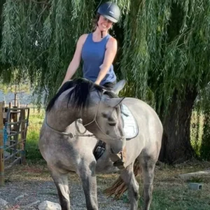 A smiling person wearing a helmet and blue sleeveless top sits on a gray horse, outdoors in front of a leafy green tree. The horse is turning its head to the left.