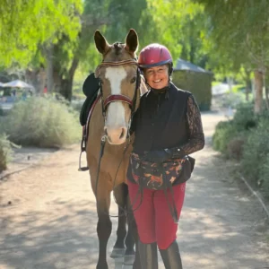 A woman wearing a red helmet and riding gear stands on a sunlit path, smiling beside a light brown horse with a white blaze, surrounded by green trees.