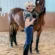 A person in riding gear stands in an indoor arena, gently holding and kissing the face of a brown horse. The horse stands calmly beside them on sandy ground near a wall marked with the letter A.
