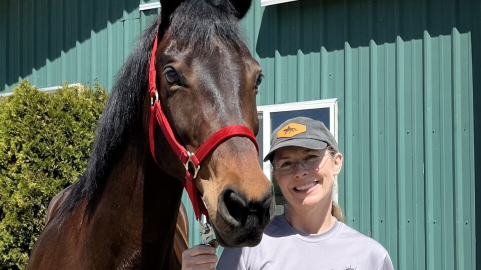 Strides Member Cindi, wearing a gray t-shirt and cap, smiles next to a brown horse in a red halter. They stand outside a green metal building on a sunny day.