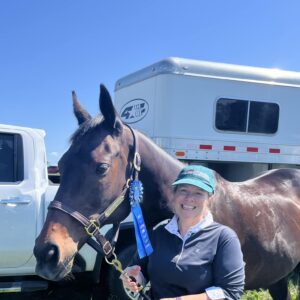 A smiling woman in a green cap and white pants stands next to a brown horse with a blue ribbon, after a 5 Day Riding Bootcamp, with a horse trailer and white truck in the background on a sunny day.