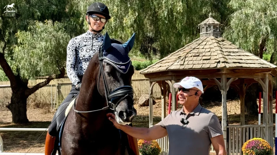 A smiling woman in riding gear sits on a dark horse, while Steffen Peters, wearing a cap and sunglasses, stands beside them, petting the horse. Behind them is a wooden gazebo and green trees—a perfect clinic recap moment.