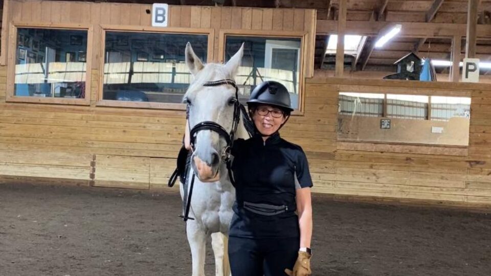 Linda, a Strides member, stands indoors next to a white horse in riding gear and helmet, smiling inside an arena with wooden walls and a large window. Riding arena letters B and P are visible on the wall behind them.
