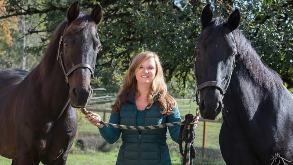 Strides member Shonnessy, a woman with long light brown hair in a teal vest, stands outdoors holding the reins of two large black horses, one on each side, with trees and a fence in the background.