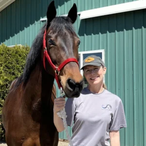 A woman in a gray T-shirt and cap smiles while standing next to a brown horse with a red halter in front of a green metal building on a sunny day.