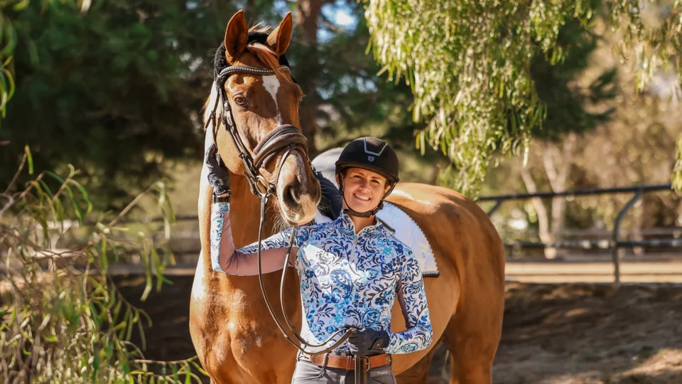 A person sporting a riding helmet, gloves, and a patterned shirt stands outdoors next to a brown horse, smiling and holding the horse’s bridle as they enjoy the sunlight—showcasing joyful Strides 2024 moments.