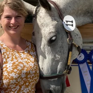 A woman in a yellow floral top smiles beside a gray horse wearing a bridle and number tag 11, with its head leaning affectionately on her. Blue and white ribbons hang in the background.