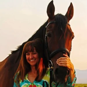 A woman with brown hair and a colorful shirt smiles while standing next to a brown horse, resting her hand on the horse’s face. The background appears to be an outdoor setting with soft evening light.