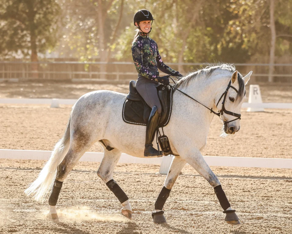 A person wearing a helmet and floral top smiles while riding a white horse in a sandy outdoor arena, surrounded by trees and sunlight. The horse is fitted with black tack and protective boots.