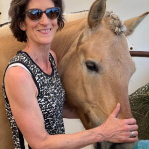 A woman wearing sunglasses and a sleeveless patterned top stands beside a light brown horse, gently holding its head. The scene appears to be outdoors near a metal fence.