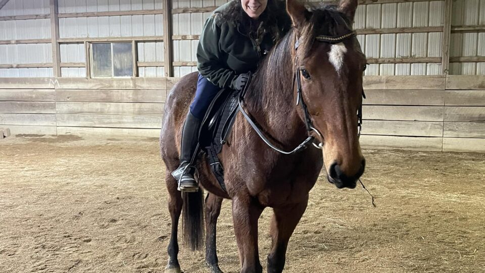 Strides member Wendi, wearing a helmet and dark clothing, is sitting and smiling on a brown horse inside a large indoor riding arena with a dirt floor and wooden walls.