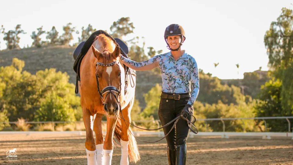 A smiling person in a helmet and riding clothes stands outdoors holding the reins of a brown horse. The fresh air, bright trees, and sunny sky create the perfect backdrop for our Spring newsletter featuring horses in action.