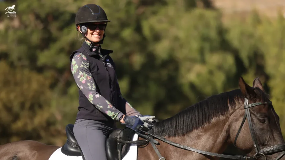A person wearing a helmet, sunglasses, and patterned long-sleeve shirt rides a dark horse outdoors, smiling and holding the reins—a moment showcasing riding improvement amid a blurred background of greenery.