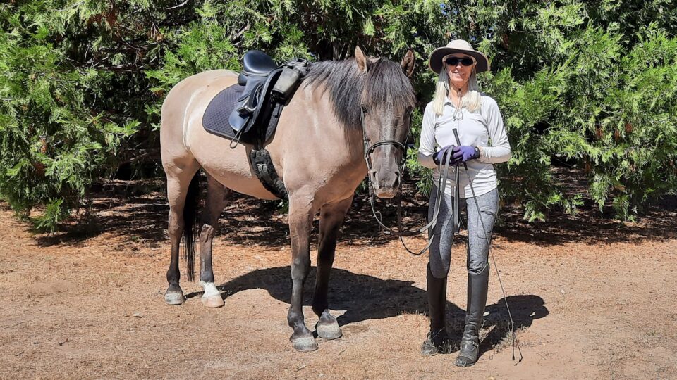 Cindy, a Strides member in riding clothes and sunglasses, stands smiling beside a saddled horse in a sunny, wooded area with green trees in the background.