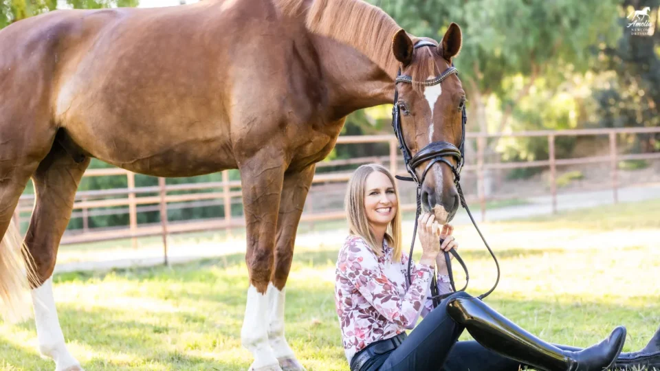 A smiling woman in riding boots sits on the grass, holding the reins of a brown horse beside her in a sunny, fenced outdoor area—a perfect Valentine scene for February 2025 with trees in the background.