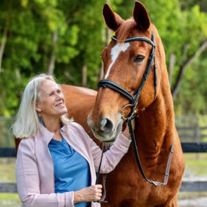 A smiling woman with long blonde hair, wearing a light pink jacket and blue shirt, stands outdoors holding the bridle of a large brown horse with a white blaze on its forehead. Green trees are visible in the background.