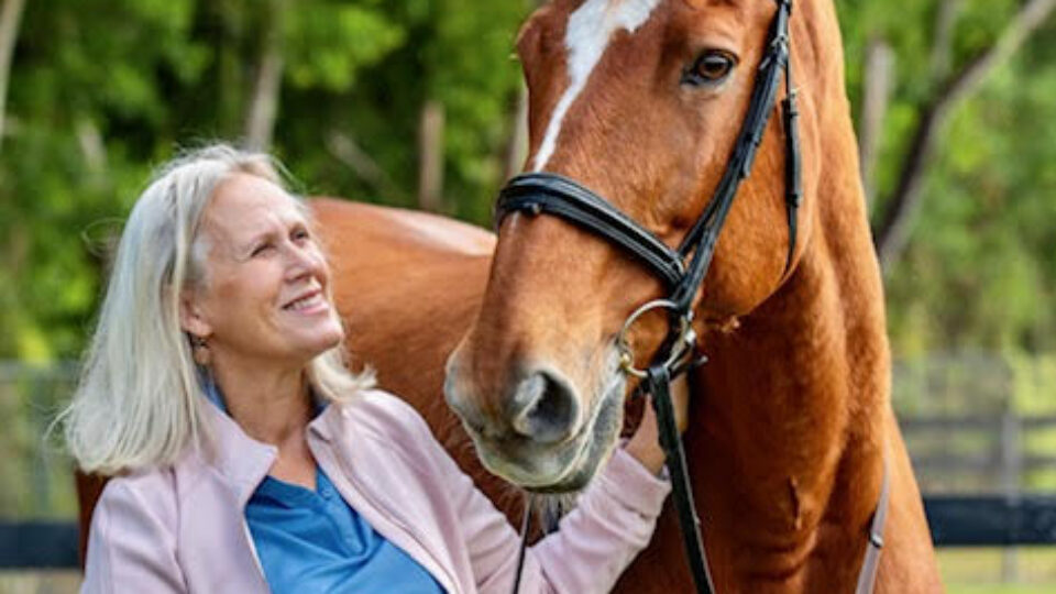 Janet, a Strides member with long blond hair, smiles as she stands outdoors in a light pink jacket and blue shirt, holding the bridle of a tall brown horse with a white blaze. Lush green trees fill the background.