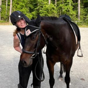A person wearing a helmet and riding gear stands next to a dark brown horse on a gravel surface, green trees behind them. Smiling, they lean close to the horse’s head, ready to take on the Sitting Trot Challenge together.