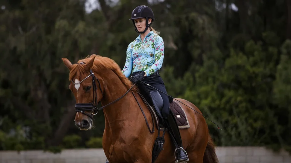 A person wearing a black helmet, colorful long-sleeve shirt, and black pants rides a chestnut horse outdoors during dressage training, with trees and greenery in the background.