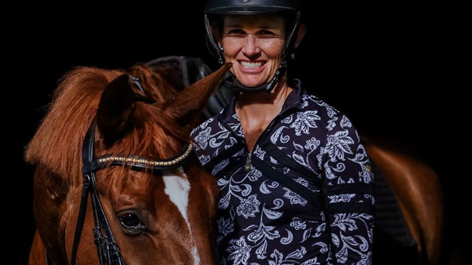 A smiling person in a patterned shirt and helmet stands beside a brown horse with a white blaze, both facing the camera against a dark background.